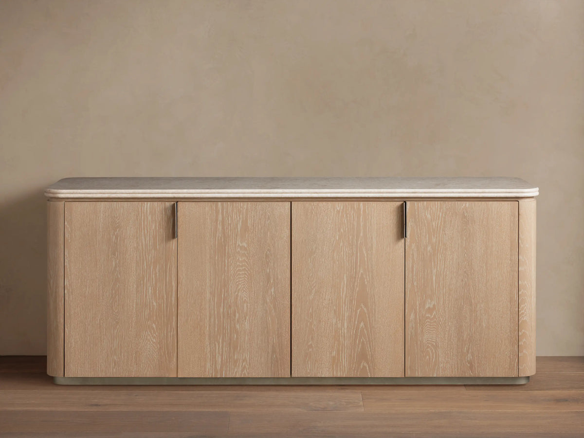 Caden Sideboard in Sand finish with light wood grain, 4 doors, brushed nickel pulls, cream stone top, on wood floor.