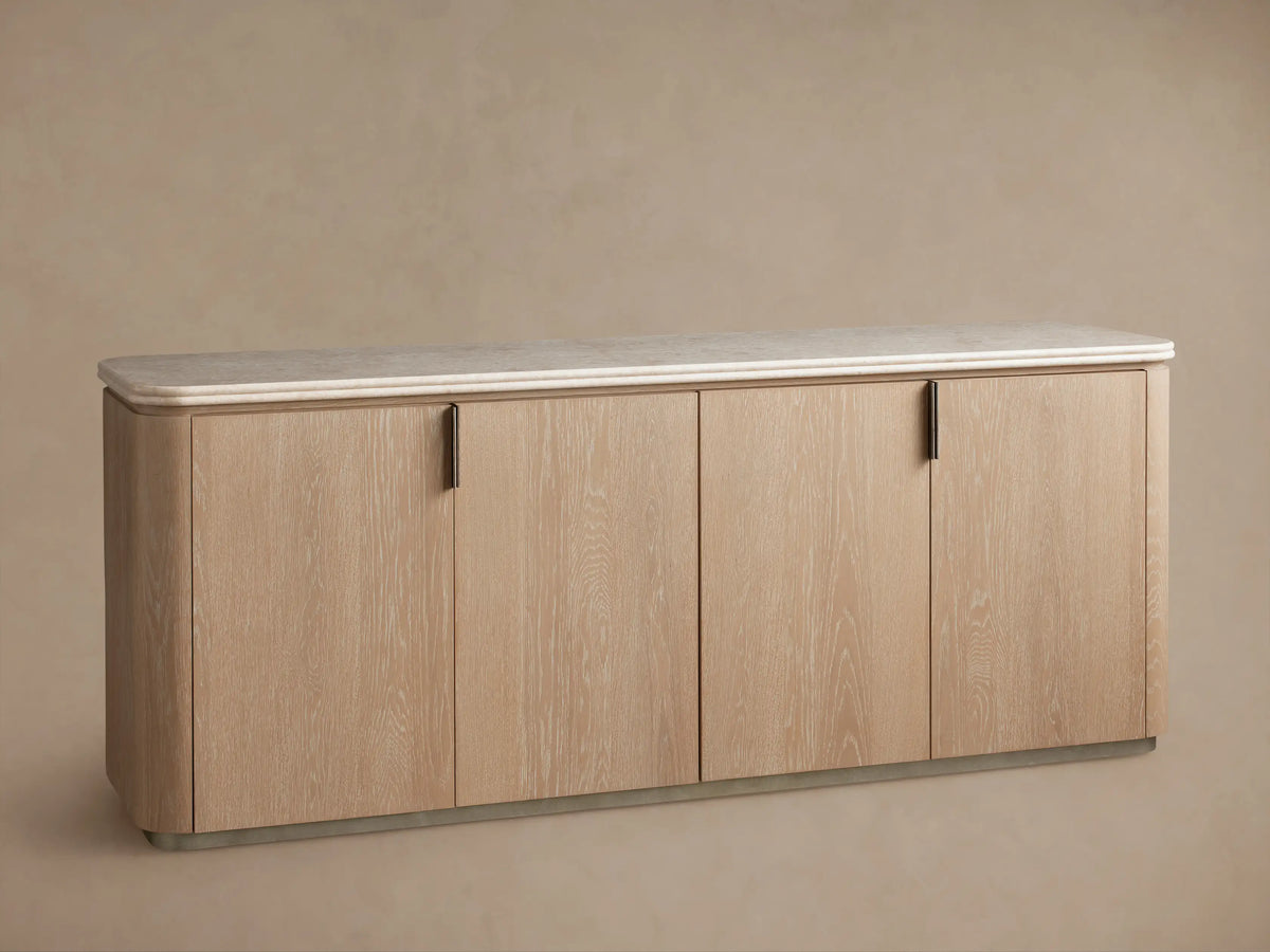 Caden Sideboard in Sand finish with light wood grain, 4 doors, brushed nickel pulls, cream stone top, on wood floor.