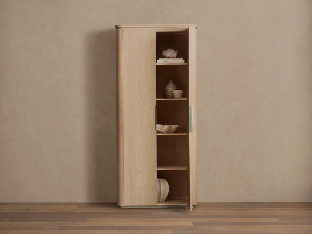 Caden Cabinet in light sand-colored wood, rounded edges, standing against a tan wall. One door slightly open, revealing shelves with decor.
