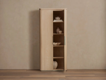 Caden Cabinet in light sand-colored wood, rounded edges, standing against a tan wall. One door slightly open, revealing shelves with decor.