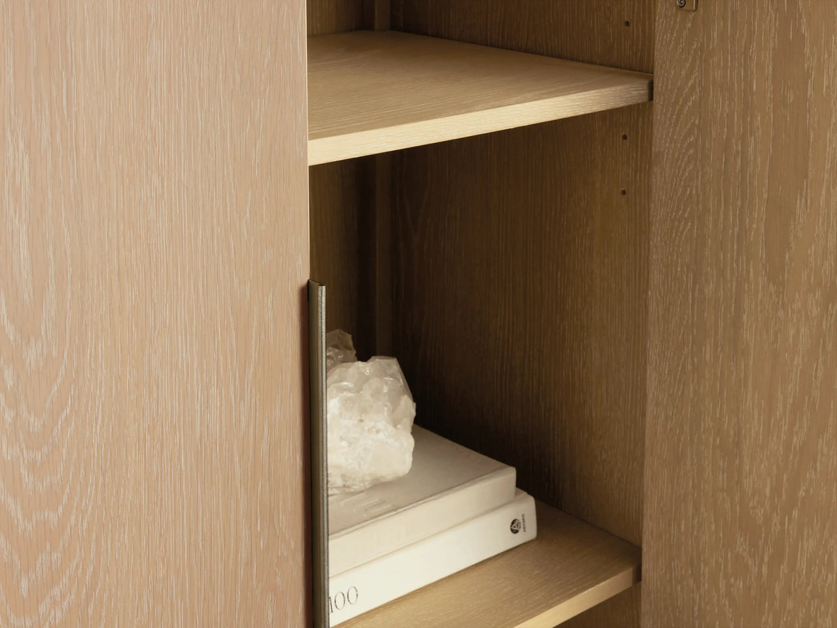 Close-up of Caden Cabinet in Sand finish, open door with brushed nickel pull, revealing wood shelves with books & crystal.