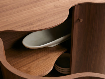 Cortina Sideboard with a wavy, contoured front design, four wooden doors, warm brown finish, and sleek metal handles.
