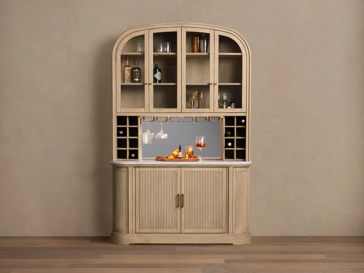 Coventry Bar: Arched light oak wood bar with glass upper doors, mirrored back, marble top, hanging glasses, and fluted lower doors.