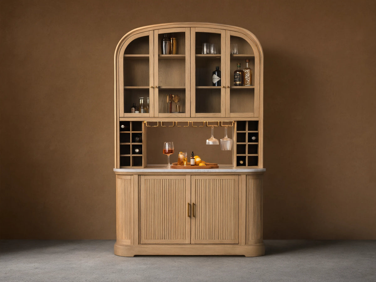 Coventry Bar: Arched light oak wood bar with glass upper doors, mirrored back, marble top, hanging glasses, and fluted lower doors.