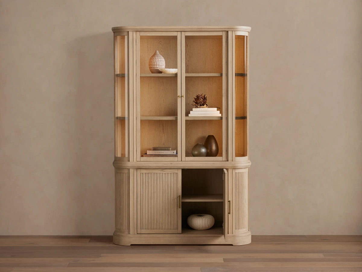 Coventry Cabinet: Light white oak display cabinet with glass top doors & illuminated shelves, one fluted bottom door ajar.