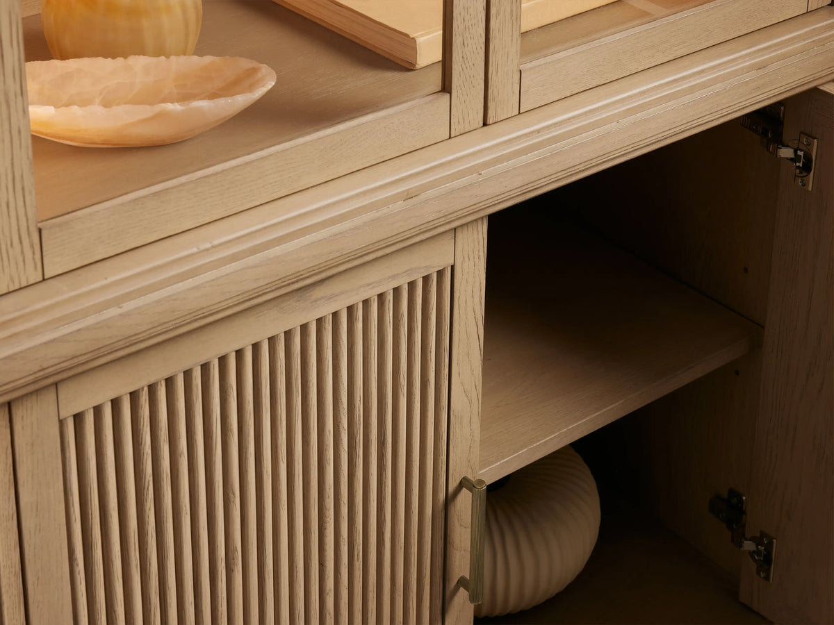 Coventry Cabinet: light white oak display cabinet with curved glass top, two shelves, and fluted wooden base doors with brass handles.