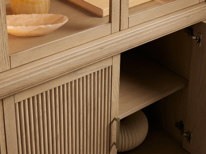 Coventry Cabinet: light white oak display cabinet with curved glass top, two shelves, and fluted wooden base doors with brass handles.