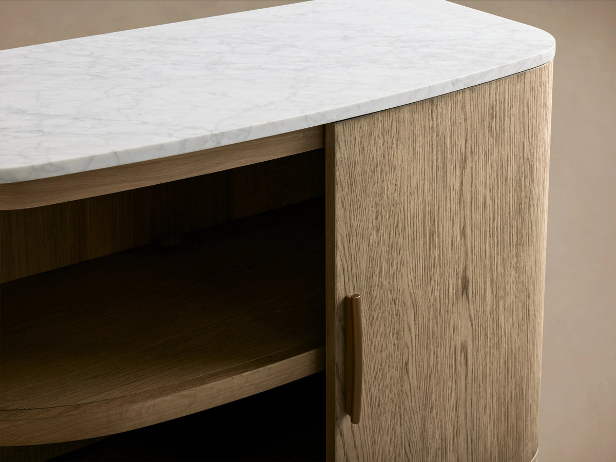 Danny Cabinet with oak wood finish, featuring a white Carrara marble top. The door is slightly open revealing an interior shelf. The handle is simple yet elegant.