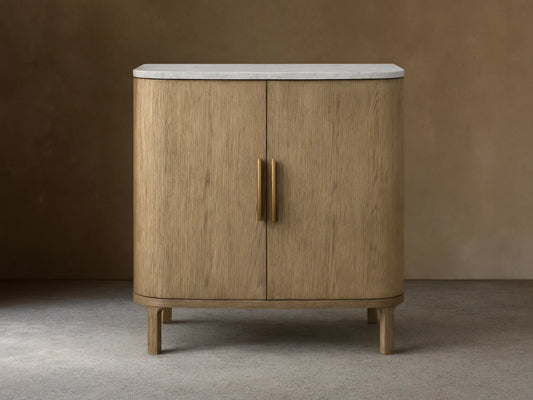 Oak Danny Cabinet with a hand-applied finish, features a smooth white Carrara marble top. Two doors with central handles, resting on short legs.