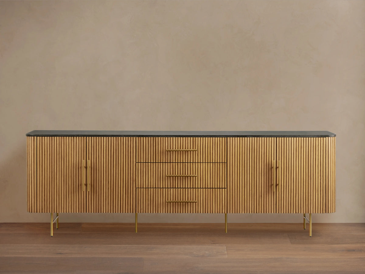 Finnley 100" Sideboard with honey finish, ribbed wood texture, dark stone top, brass hardware, and metal legs, against a beige wall.