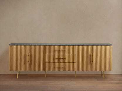 Finnley 100" Sideboard with honey finish, ribbed wood texture, dark stone top, brass hardware, and metal legs, against a beige wall.