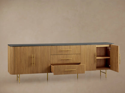 Finnley 100" Sideboard with honey finish, ribbed wood texture, dark stone top, brass hardware, and metal legs, against a beige wall.