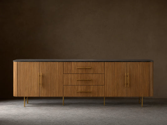 Finnley 100" Sideboard with honey finish, ribbed wood texture, dark stone top, brass hardware, and metal legs, against a beige wall.