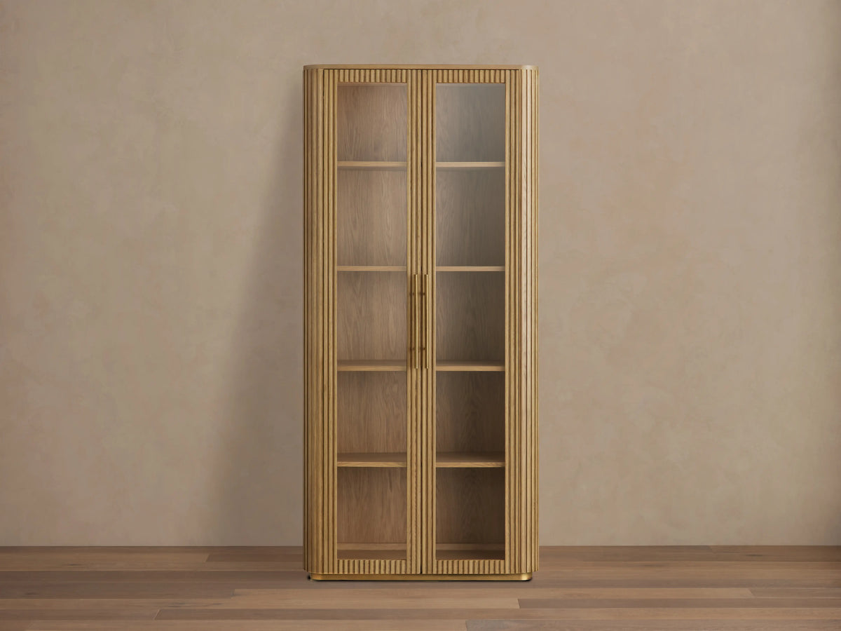 Finnley Glass Cabinet with honey-finished oak wood, two glass doors, brass handles, and four wooden shelves against a beige wall.