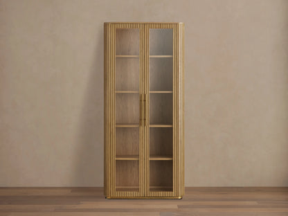 Finnley Glass Cabinet with honey-finished oak wood, two glass doors, brass handles, and four wooden shelves against a beige wall.