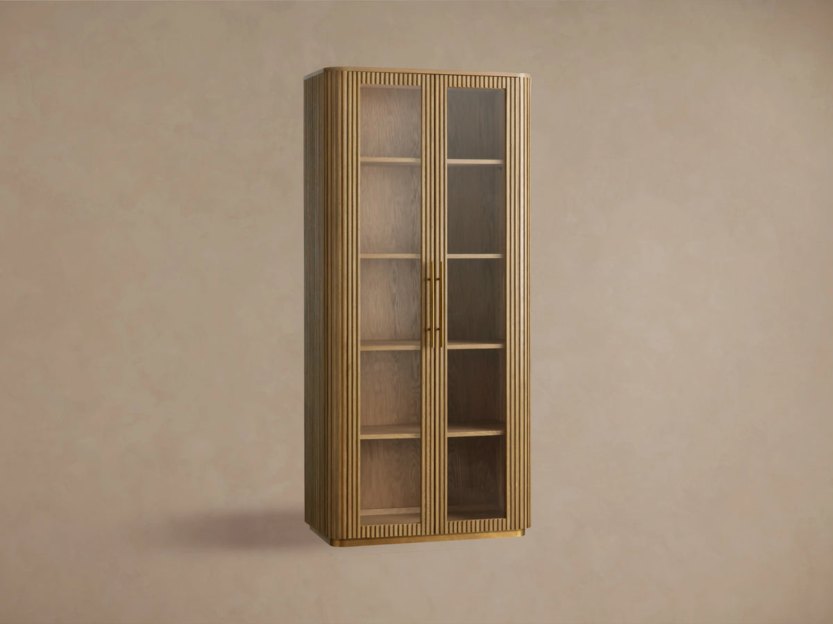 Finnley Glass Cabinet with honey-finished oak wood, two glass doors, brass handles, and four wooden shelves against a beige wall.