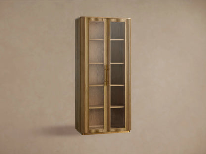 Finnley Glass Cabinet with honey-finished oak wood, two glass doors, brass handles, and four wooden shelves against a beige wall.