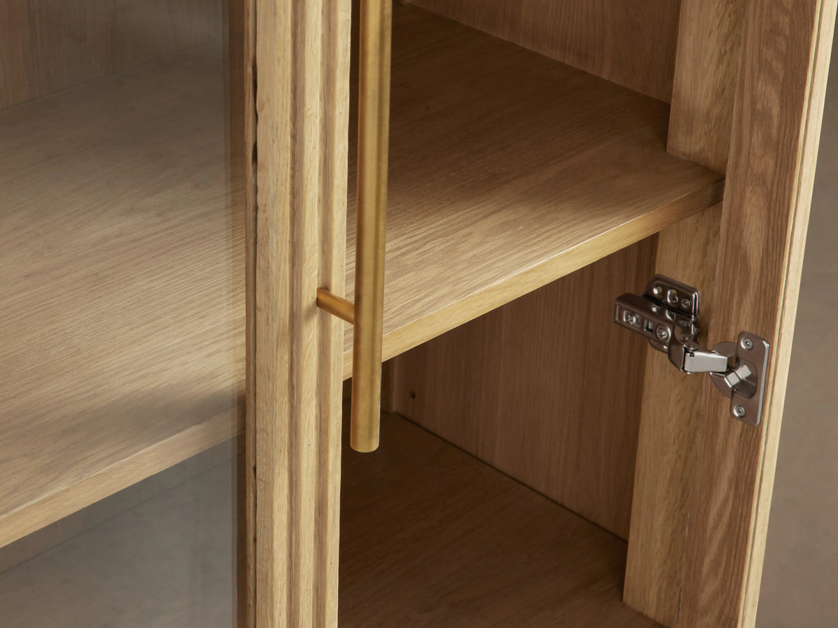 Finnley Glass Cabinet with honey finish, featuring two paneled tempered glass doors, brass pulls, and visible shelves inside.