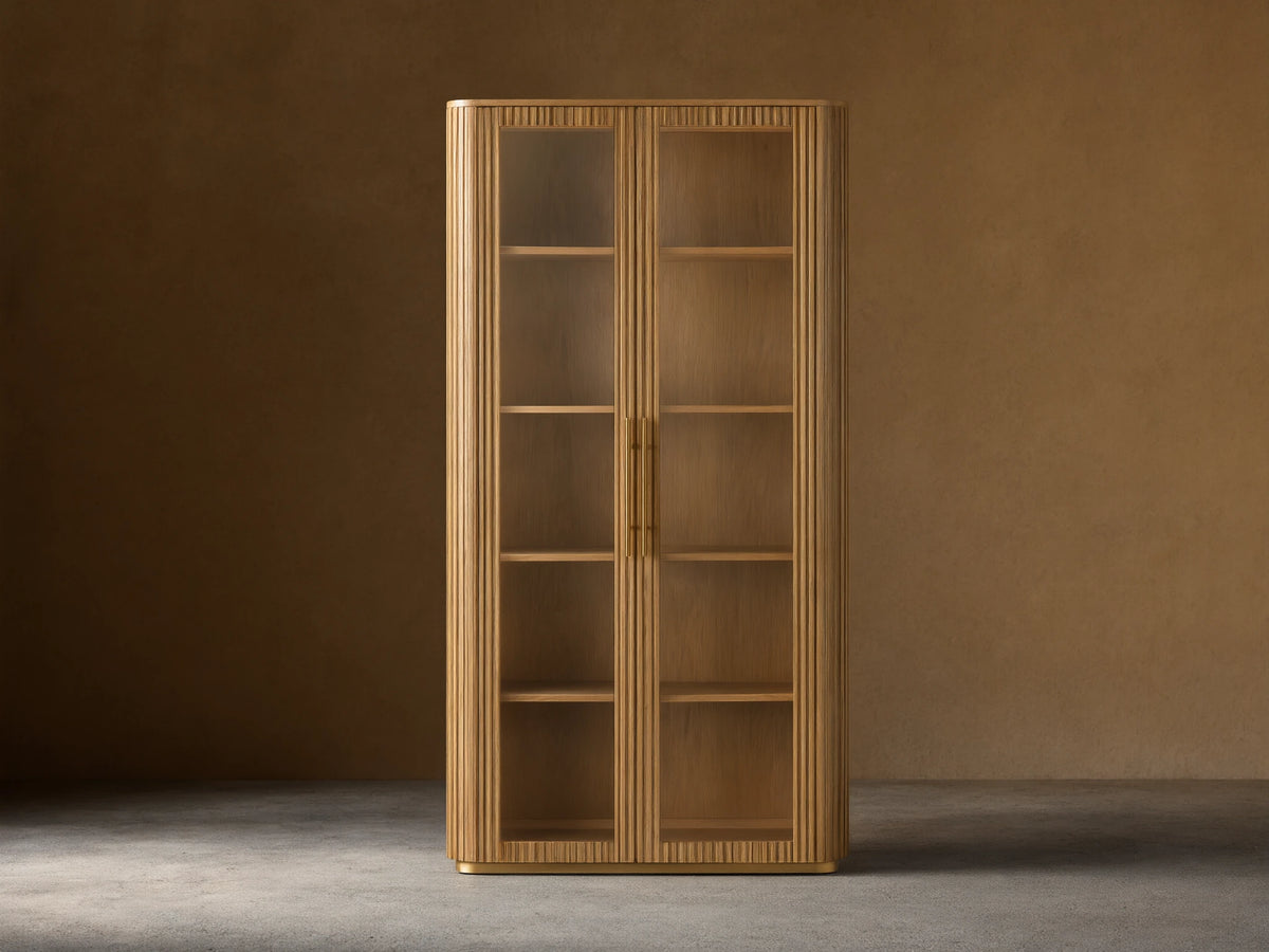 Finnley Glass Cabinet with honey-finished oak wood, two glass doors, brass handles, and four wooden shelves against a beige wall.