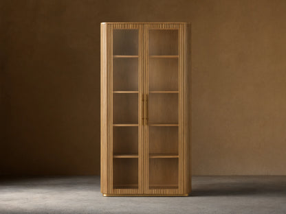 Finnley Glass Cabinet with honey-finished oak wood, two glass doors, brass handles, and four wooden shelves against a beige wall.