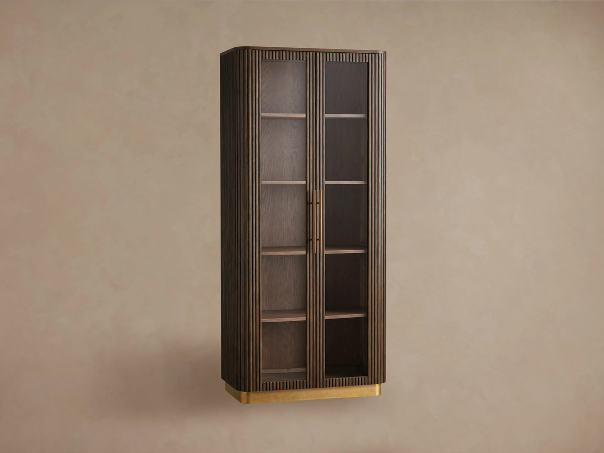 Finnley Glass Cabinet: dark brown wood, two glass doors, brass pulls & plinth base, fluted sides, on wood floor against a beige wall.