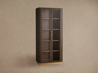 Finnley Glass Cabinet: dark brown wood, two glass doors, brass pulls & plinth base, fluted sides, on wood floor against a beige wall.
