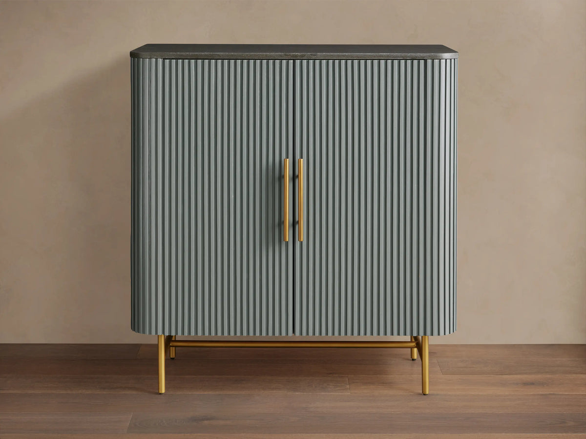 Finnley Bar Cabinet in pumice with vertical reeding detail, dark top, and gold brass-finished handles & legs.