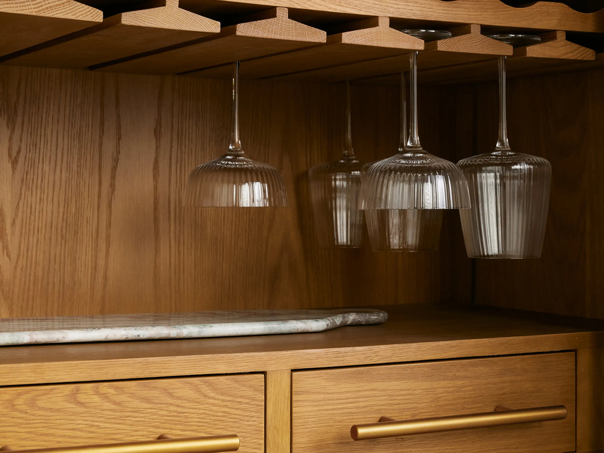 Finnley Bar Cabinet with dark stone top over a light wood pull-out shelf. Fluted glass with drink, stirrer, green napkin, dried orange.