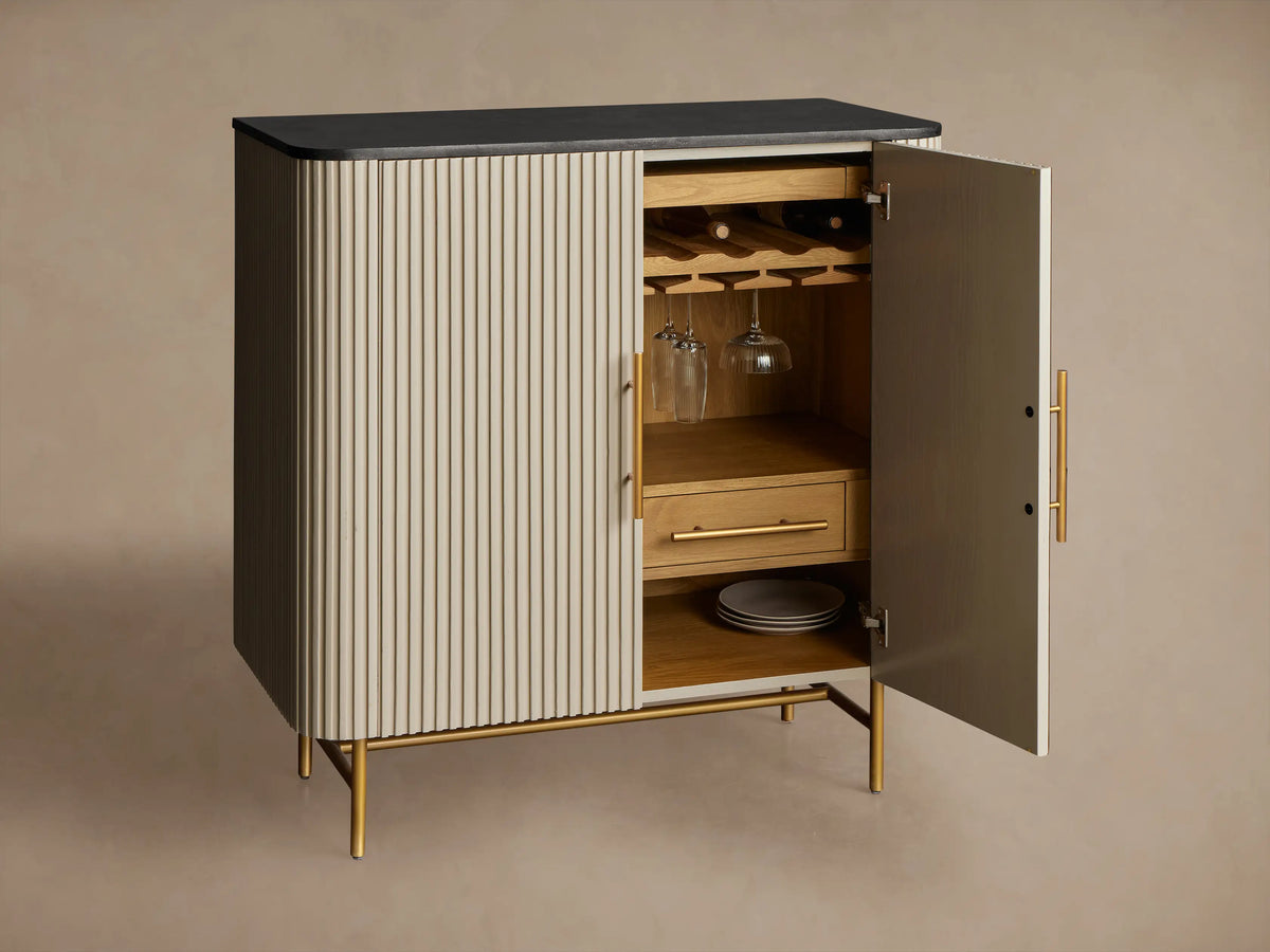 Finnley Bar Cabinet with dark top, reeded light-colored doors, vertical brass handles, and thin brass legs on wood floor.
