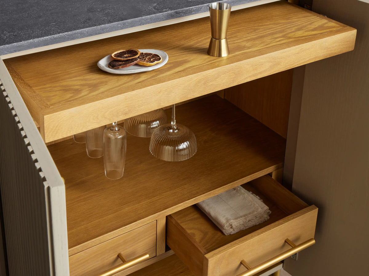 Finnley Bar Cabinet, pumice reeded front, dark bluestone top, brass legs. One door open revealing wine, glass, & plate storage.