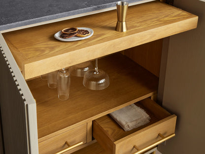 Finnley Bar Cabinet, pumice reeded front, dark bluestone top, brass legs. One door open revealing wine, glass, & plate storage.