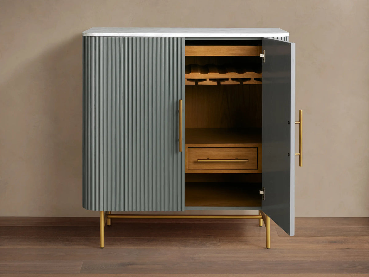 Finnley Bar Cabinet: Pumice-colored reeded doors, white marble top, brass handles, & gold legs, centered on wooden floor.