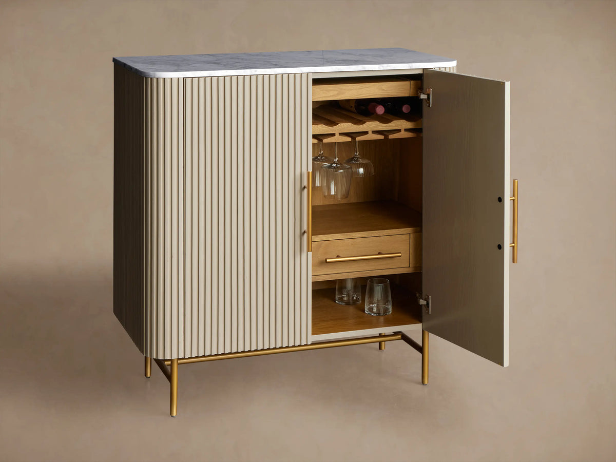 Finnley Bar Cabinet, pumice finish with vertically reeded doors, white marble top, thin brass handles & legs, front view.