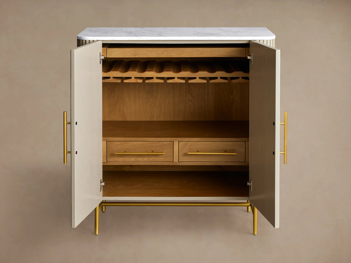 Close-up of open Finnley Bar Cabinet, showing reeded Pumice door, brass handles, hanging wine glasses, wine bottles, and drawer.