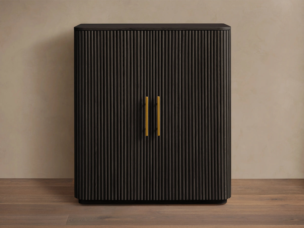 Finnley Wood Bar Cabinet with a dark ebony finish, vertical grooves, and two vertical gold handles. It stands on a wooden floor against a beige wall.