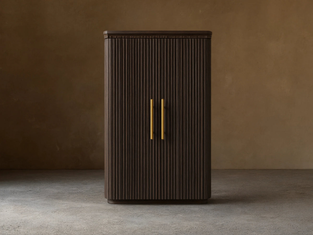 Finnley Wood Bar Cabinet with a dark ebony finish, vertical grooves, and two vertical gold handles. It stands on a wooden floor against a beige wall.