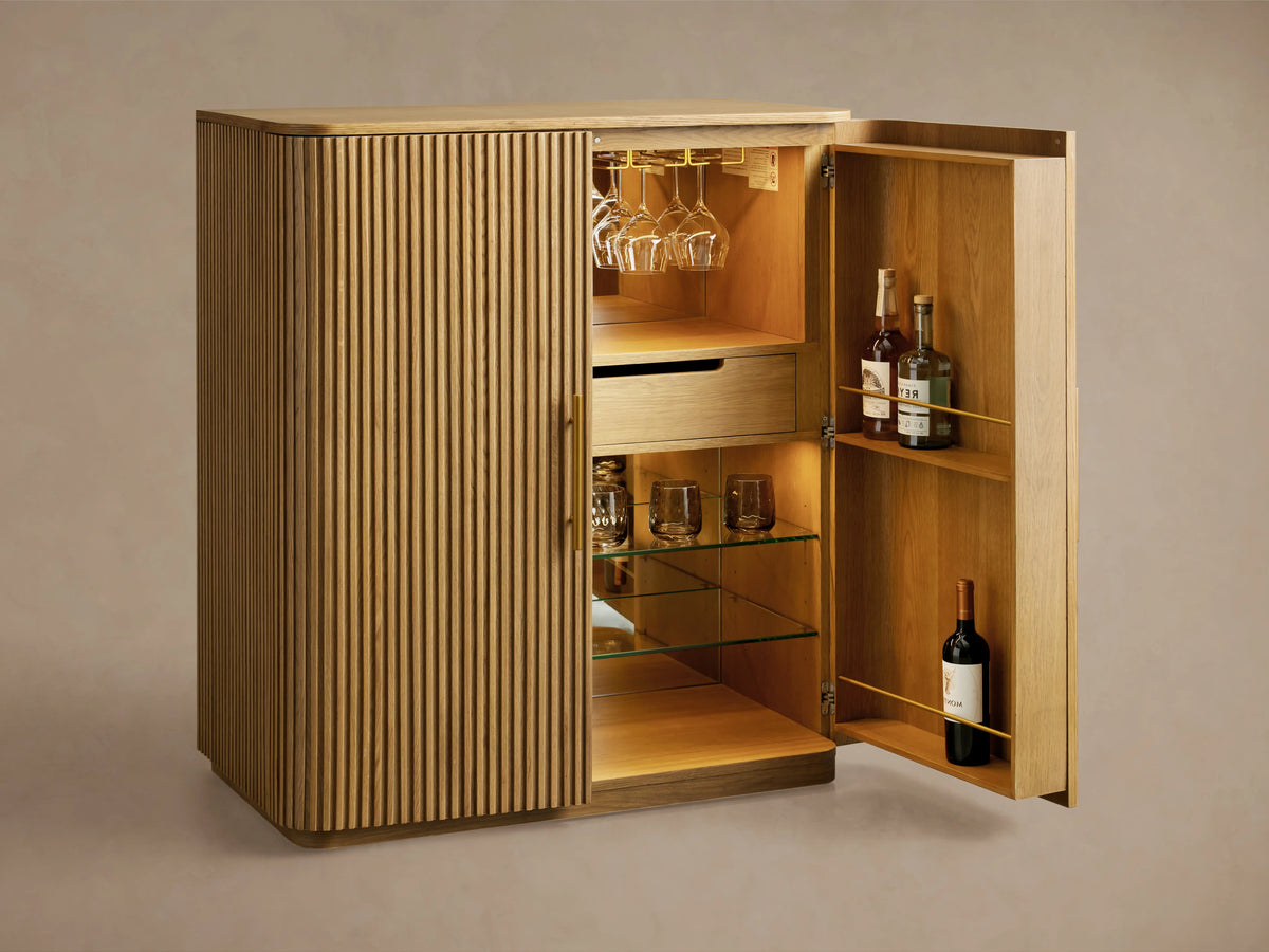 Finnley Wood Bar Cabinet with a dark ebony finish, vertical grooves, and two vertical gold handles. It stands on a wooden floor against a beige wall.