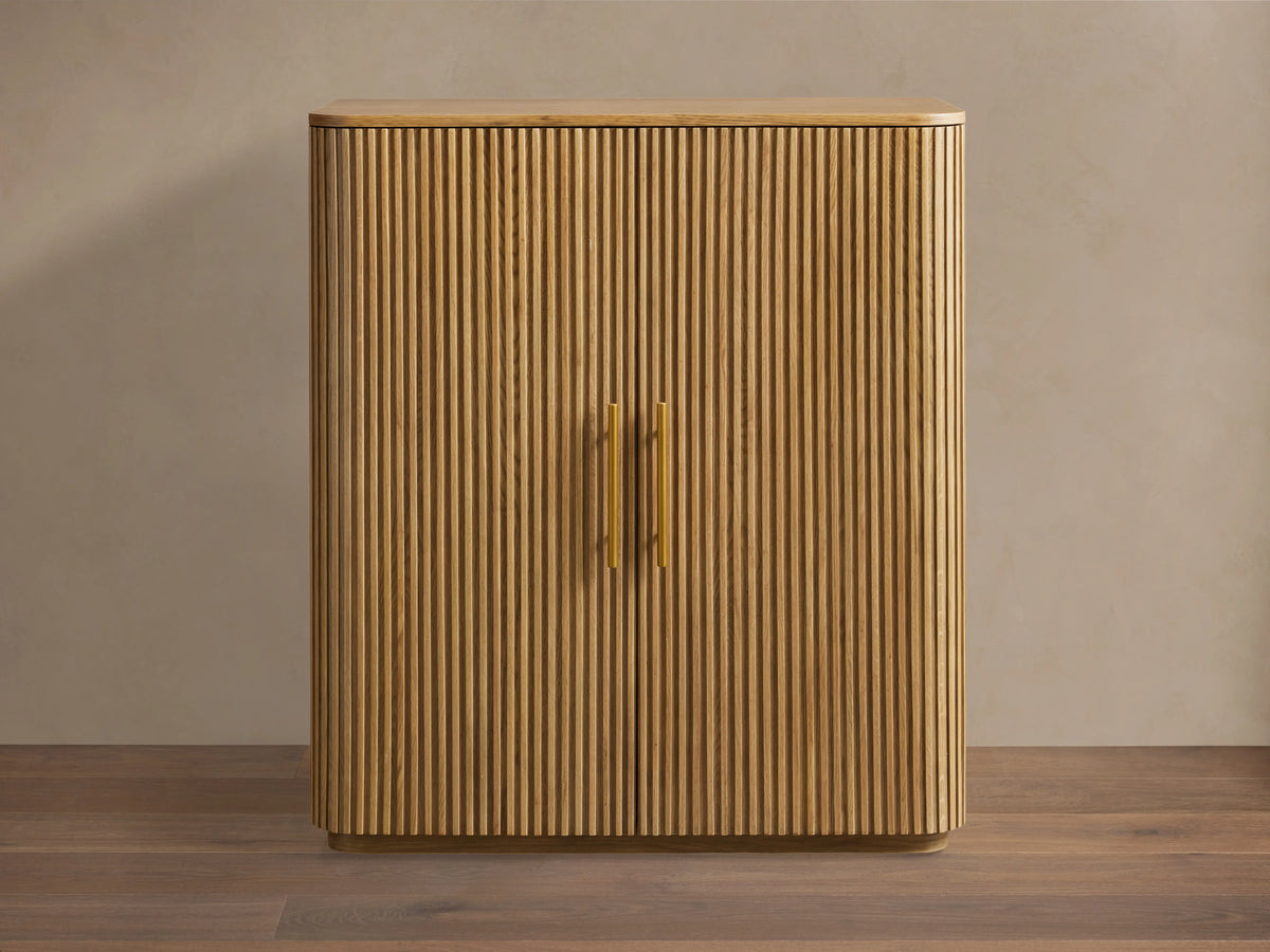 Finnley Wood Bar Cabinet with a honey finish, featuring vertical ribbed design, two central gold handles, and curved edges.