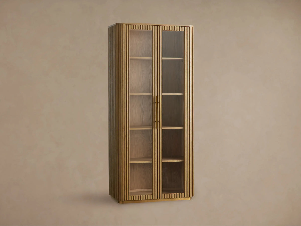 Finnley Glass Cabinet. Tall, honey-finished wood cabinet with vertical fluting, two glass doors, brass pulls, and interior shelves.