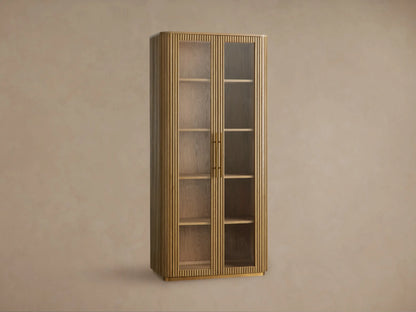 Finnley Glass Cabinet. Tall, honey-finished wood cabinet with vertical fluting, two glass doors, brass pulls, and interior shelves.