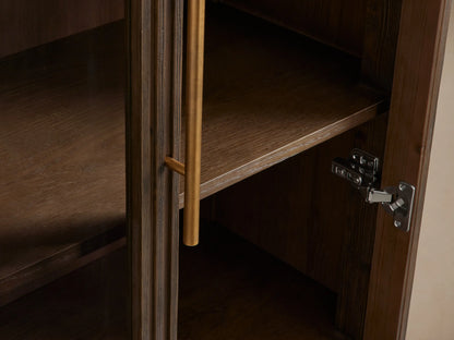 Finnley Glass Cabinet, dark mocha wood with fluted details, two glass doors, brass handles & base, against a beige wall.