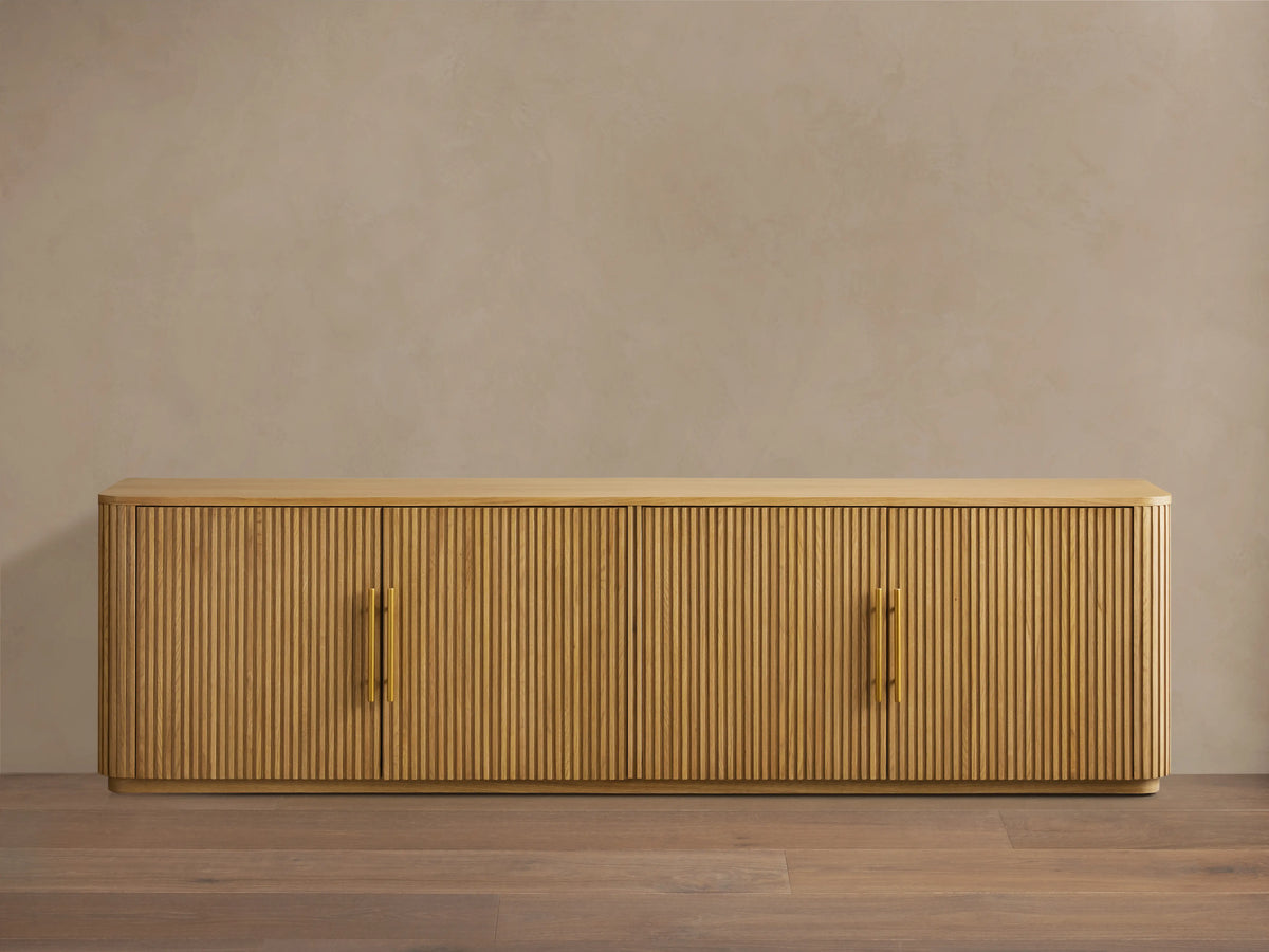 Finnley Wood Media Console with a ribbed front design in Honey finish, featuring brass-finished handles, set against a neutral wall.