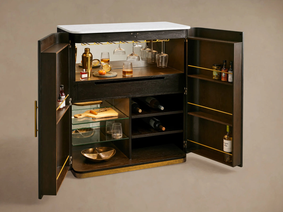 Finnley Plinth Bar Cabinet with dark fluted doors, brass handles & plinth base, and a white marble top against a beige wall.