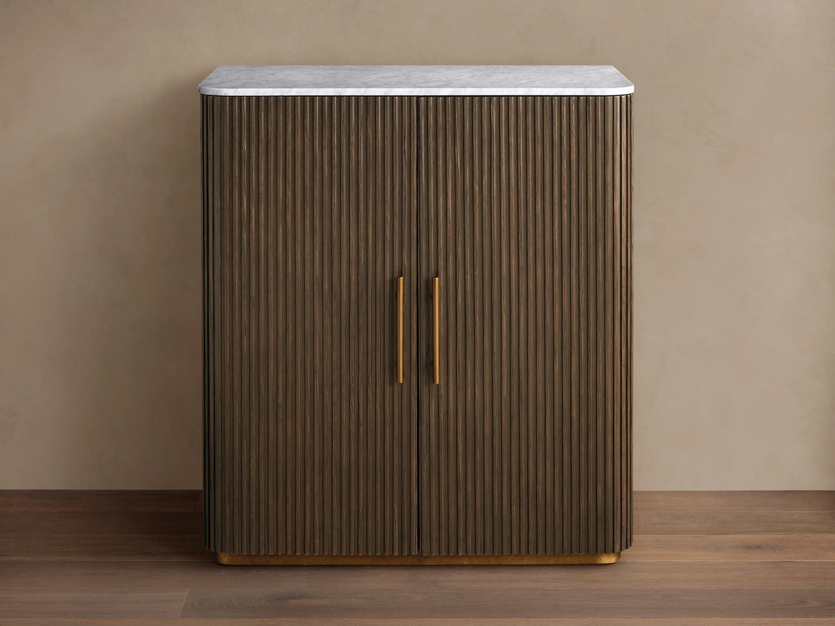 Finnley Plinth Bar Cabinet with dark fluted wood doors, white marble top, brass handles, and plinth base.
