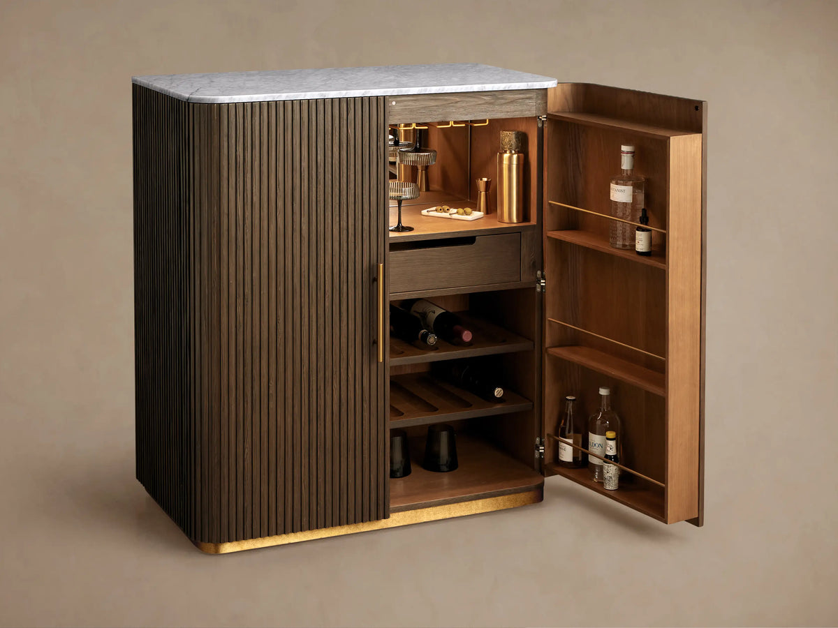 Finnley Plinth Bar Cabinet with dark fluted wood doors, white marble top, brass handles, and plinth base.