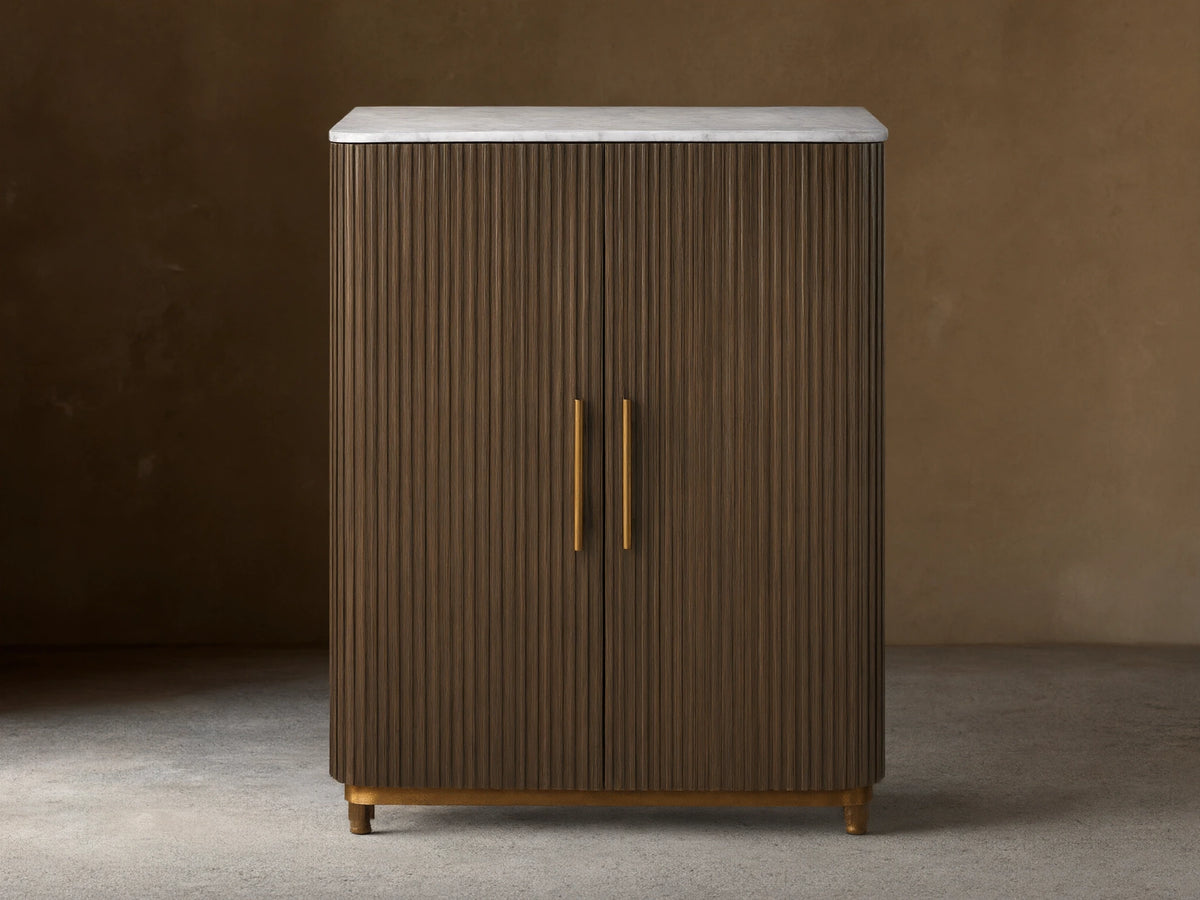 Finnley Plinth Bar Cabinet with dark fluted wood doors, white marble top, brass handles, and plinth base.