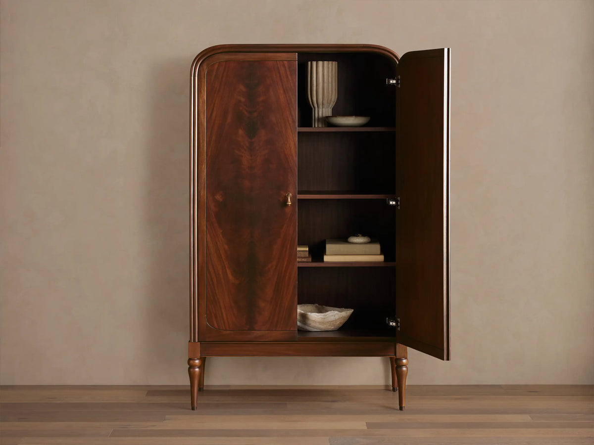 Frances Cabinet: Dark mahogany cabinet with book-matched figured veneer, rounded top, brass handles, turned legs, on a wood floor.