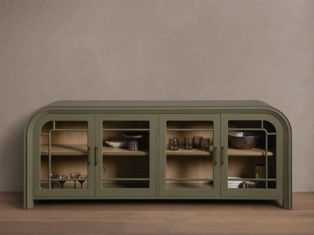 Front view of a green Gatlin Sideboard with four tempered glass doors, rounded ends, and antique brass pulls, displaying items on wood shelves.