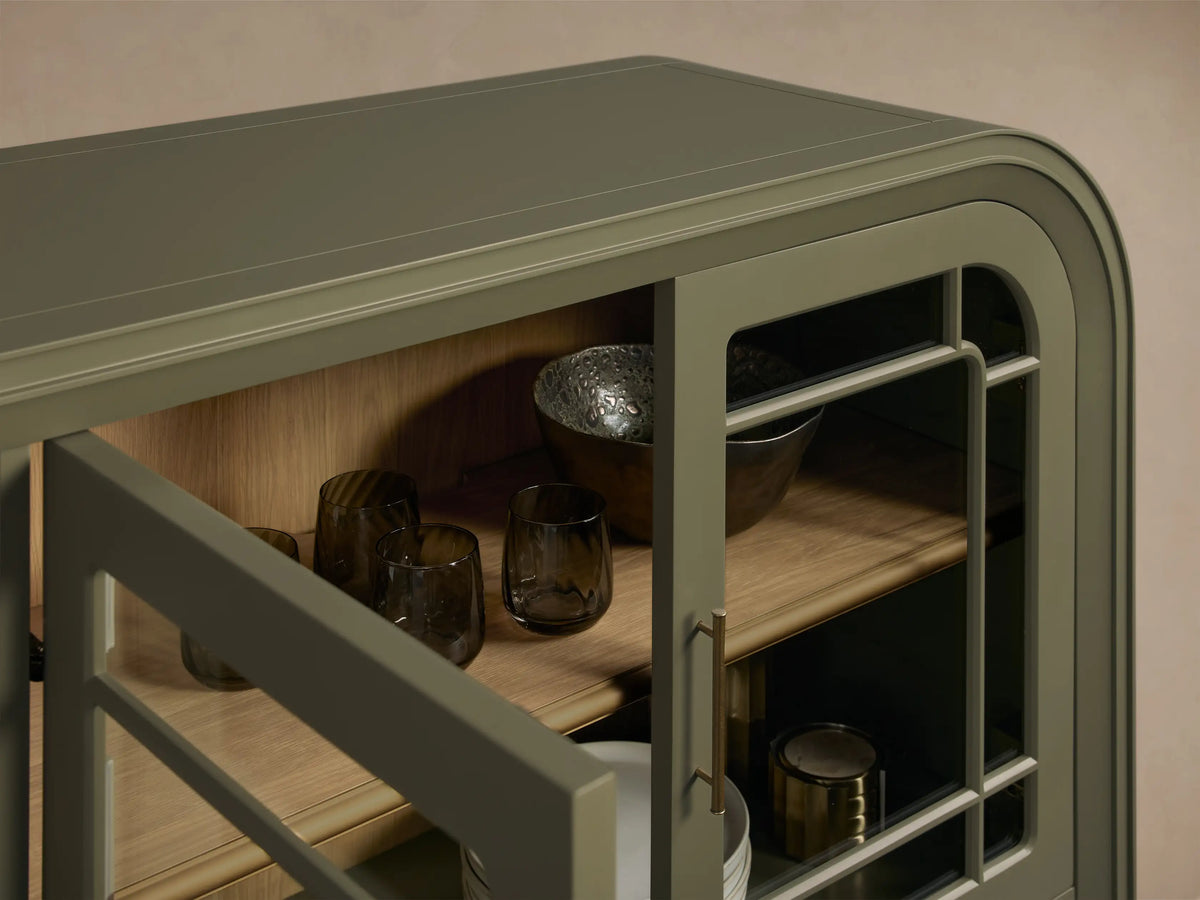 Front view of the Eclipse Gatlin Sideboard, a dark green cabinet with rounded ends, four glass doors, brass pulls, and interior wooden shelves.