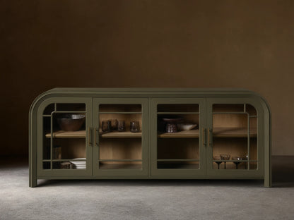 Front view of a green Gatlin Sideboard with four tempered glass doors, rounded ends, and antique brass pulls, displaying items on wood shelves.